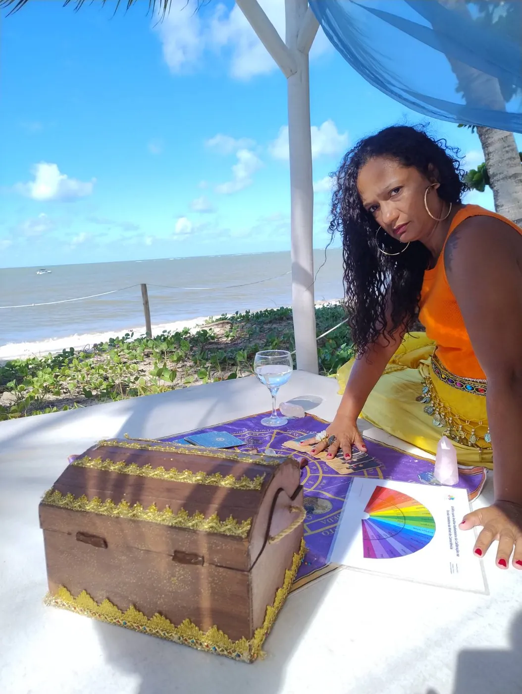 Liviã sentada à mesa de atendimento na praia, com o baú de madeira e as cartas